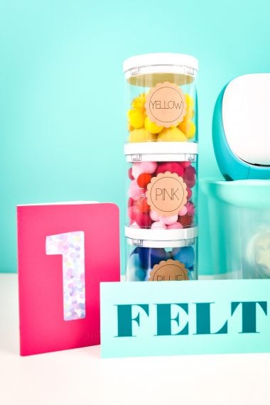 A Cricut Joy sitting on top of a clear bin, three containers with pom poms in them and labeled yellow, pink and blue, a pink card with the number 1 on it and a label that says 'Felt'.