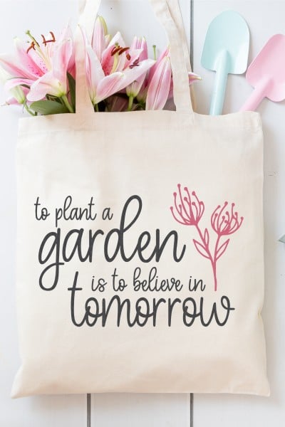 A canvas bag filled with pink lilies and two small gardening shovels in blue and pink.  The canvas bag has a picture of a flower on it with the saying, "To plant a garden is to believe in tomorrow".