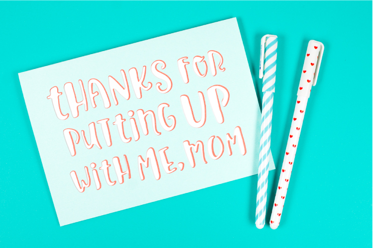 Two pens sitting next to a greeting card that says, \"Thanks for Putting Up with Me Mom\"