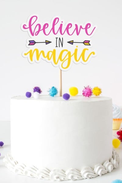 A cake decorated with white icing and colorful pom-poms and an inspirational sign saying "Believe in Magic" stuck into the top of the cake