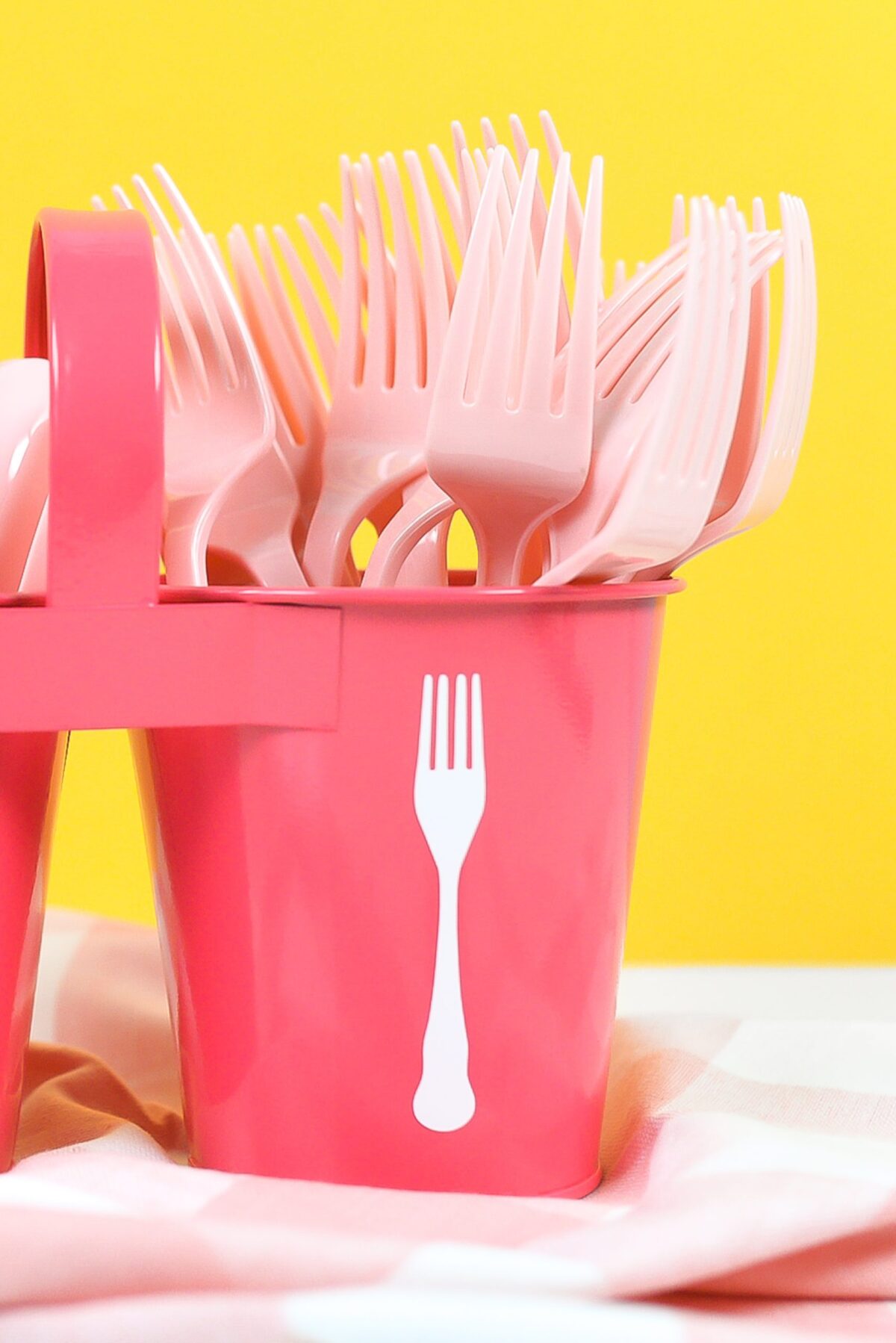 Zoomed in shot of pink utensil caddy with white spoon and for decals, filled with pink plastic forks and spoons on a yellow background.