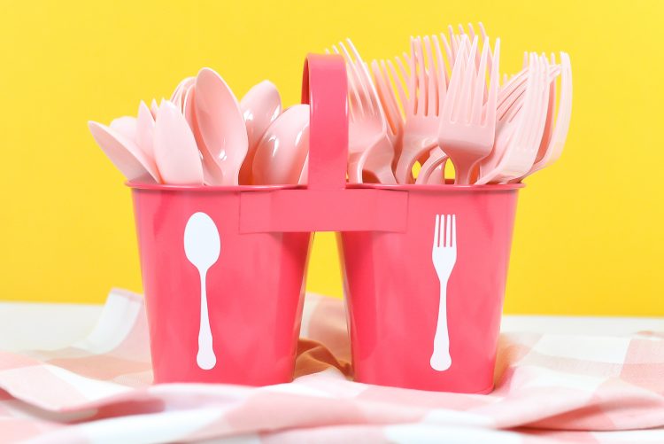 Pink utensil caddy with white spoon and for decals, filled with pink plastic forks and spoons on a yellow background.