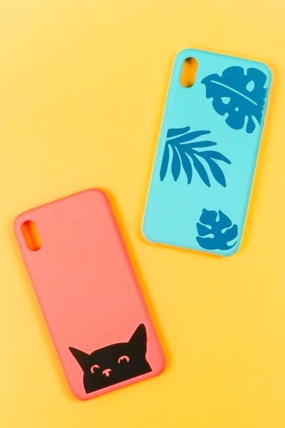Two phone cases decorated in vinyl.  One is coral colored with a black cat's head peaking out from the bottom of the case and the other is an aqua blue case with blue colored tropical leaves attached to it