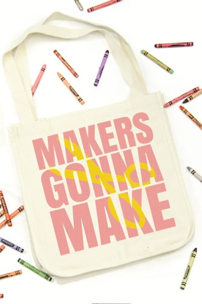 Canvas tote displaying a block knockout design using the quote "Makers Gonna Make" in pink vinyl and a yellow scissors over the top of the quote. Crayons of many colors scattered around the tote.