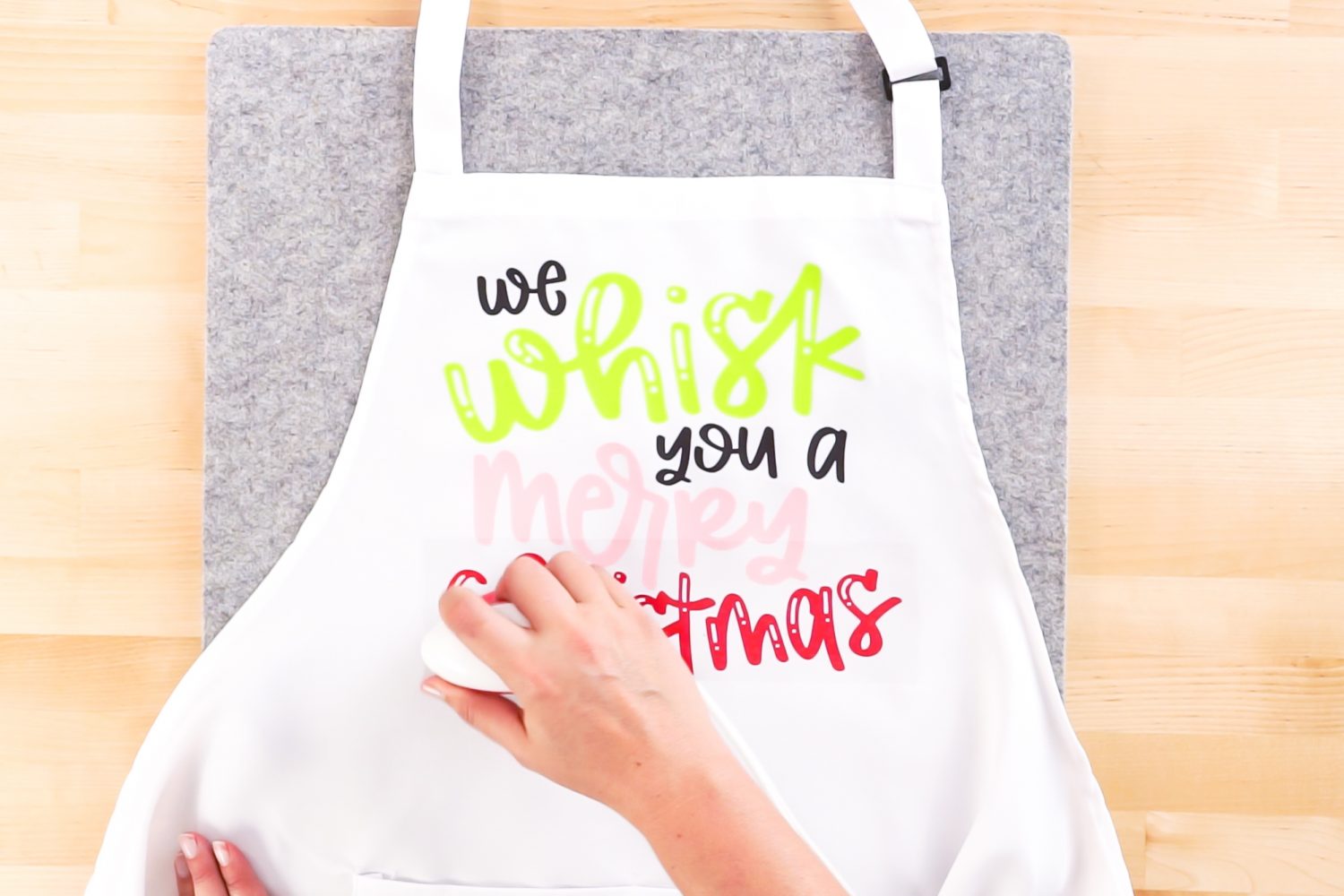 Close up picture of a person using an EasyPress to adhere vinyl to a Christmas Apron with quote on apron saying \"We whisk you a Merry Christmas\"