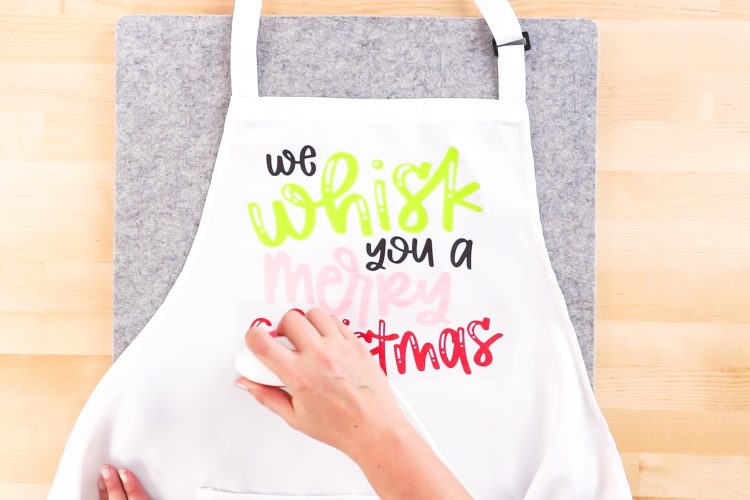 Close up picture of a person using an EasyPress to adhere vinyl to a Christmas Apron with quote on apron saying "We whisk you a Merry Christmas"