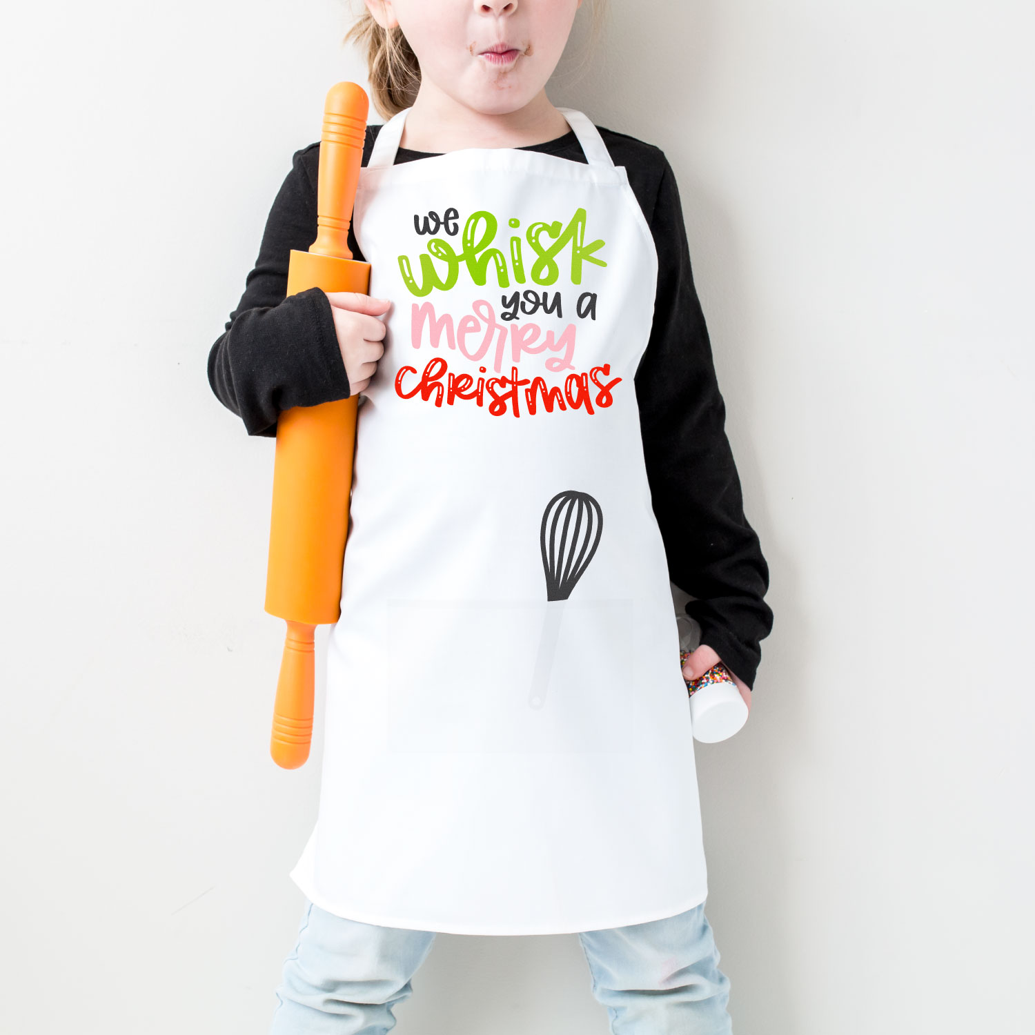 A little girl holding a rolling pin and wearing a Christmas Apron with a whisk in the apron pocket with quote on apron saying \"We whisk you a Merry Christmas\"