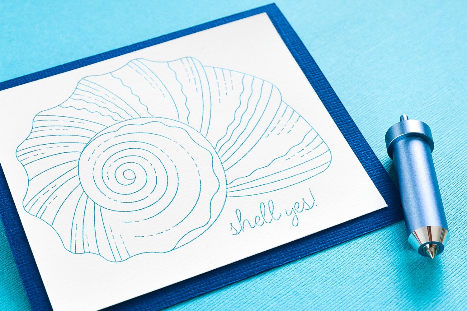 A piece of blue cardstock with a front cover of a sea shell on white cardstock that is outlined in aqua colored foil and says \"Shell yes\".  A foil tip housing unit in light blue lays next to the cardstock image