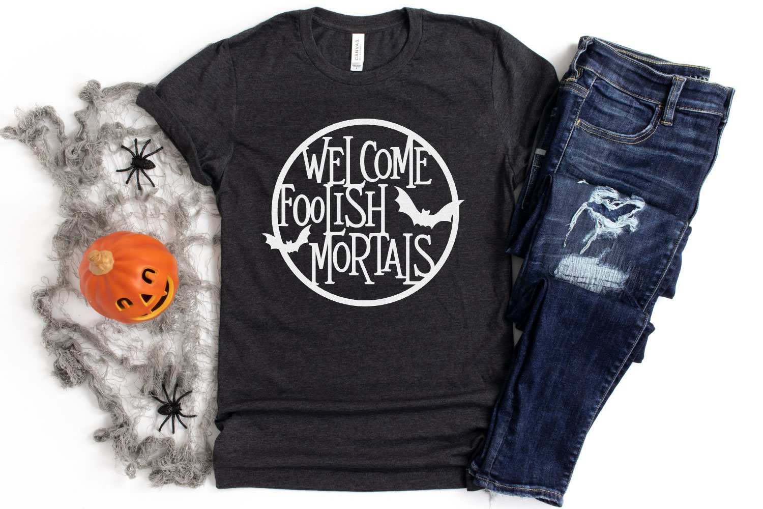 Image of a black t-shirt with quote in white vinyl stating \"Welcome Foolish Mortals\" encased within a white vinyl cirlce and two white bats.  A pair of blue jeans and some Halloween decor are next to the t-shirt