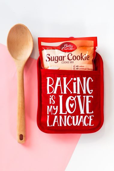 A red potholder with the quote "Baking is my Love Language" on it and a package of sugar cookie mix tucked inside the holder with a wooden spoon next to it
