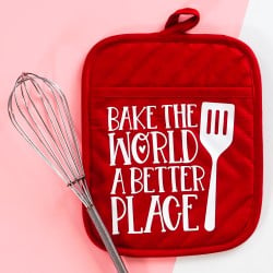 A red potholder with the quote "Bake the World a Better Place" with a spatula and whisk next to it