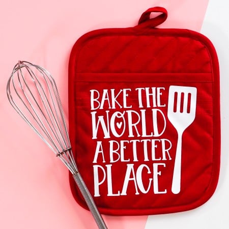 A red potholder with the quote "Bake the World a Better Place" with a spatula and whisk next to it