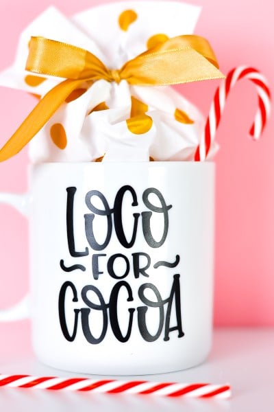 Picture of a white mug with the quote "Loco for Cocoa" in black vinyl on it.  Mug contains a red and white striped candy cane and a small package wrapped in white and gold.