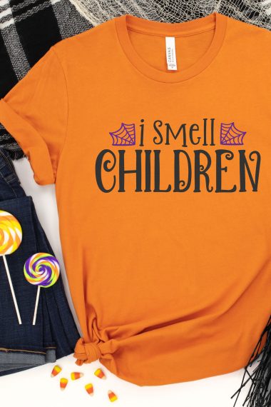 An orange t-shirt with the quote of "I Smell Children" on it along with two spider webs in purple. Two suckers lay next to the shirt.