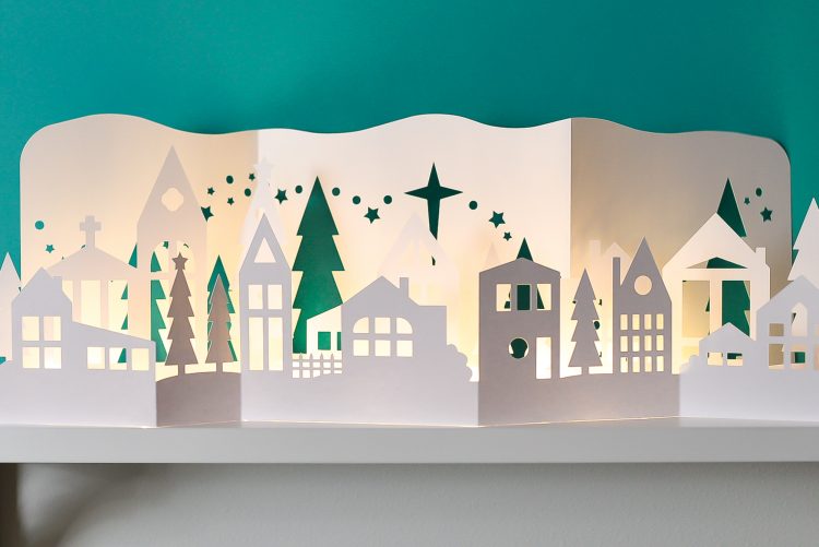 Close up image of Papercut Christmas Village on a shelf illuminated with fairy lights