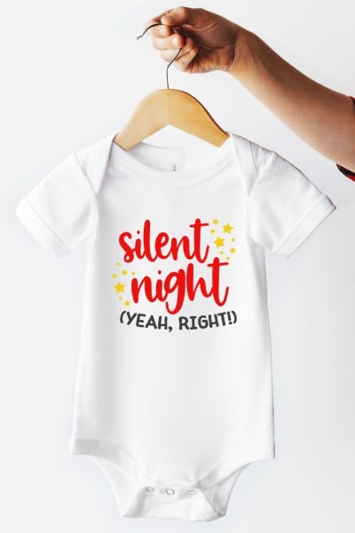 A white onesie on a hanger that is decorated with yellow stars and the words "Silent Night (yeah, right!)" on it