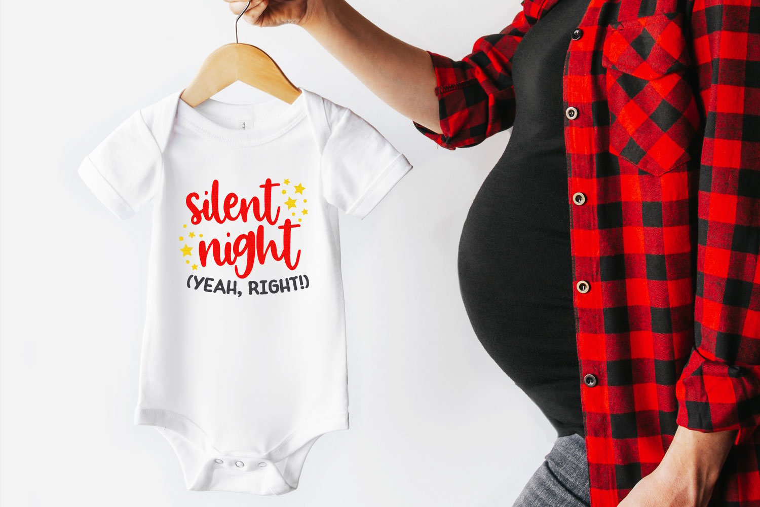 A hanger with a white onesie on it is being held by a pregnant woman in a black maternity top and a red and black buffalo checkered shirt.  The onesie is decorated with yellow stars and the words \"Silent Night (yeah, right!)\" on it 