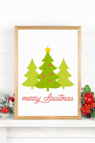 Sign in wooden frame with three green colored trees and the words Merry Christmas below the trees.  Frame is placed among holiday decorations.