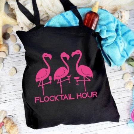 Three pink flamingo images on a black tote back with the saying Flocktail Hour