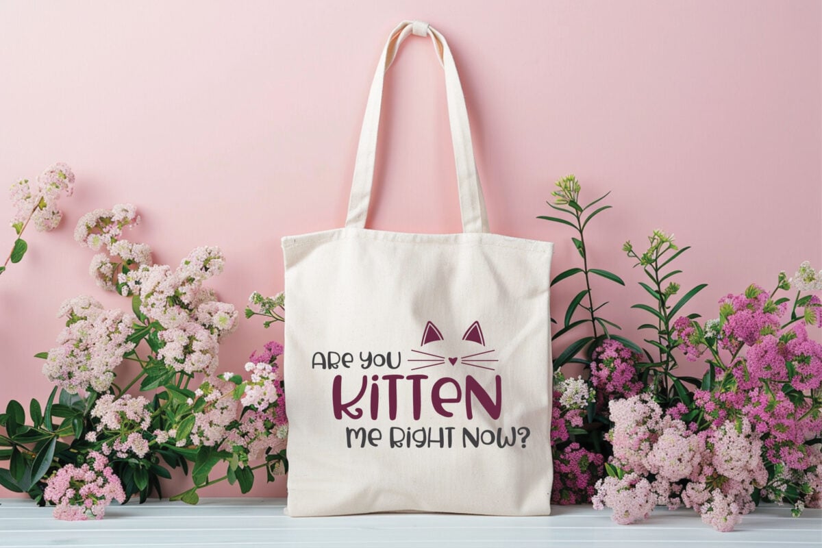 Kitten Me SVG file on a natural tote bag surrounded by purple wildflowers