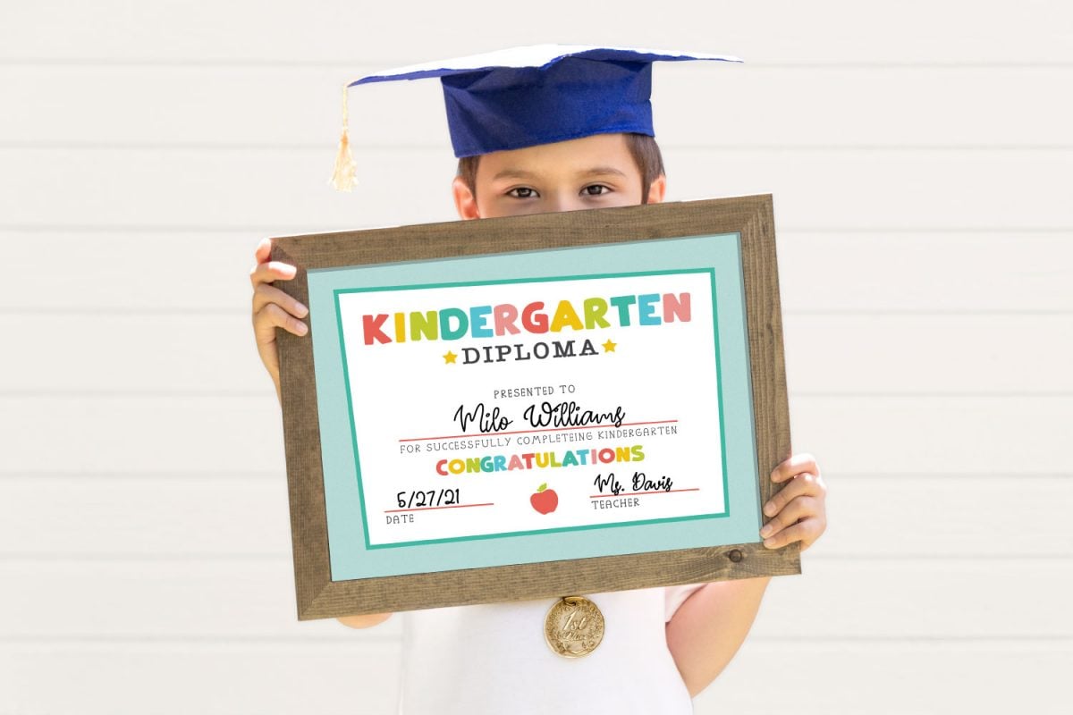 Child holding framed kindergarten diploma while wearing grad cap