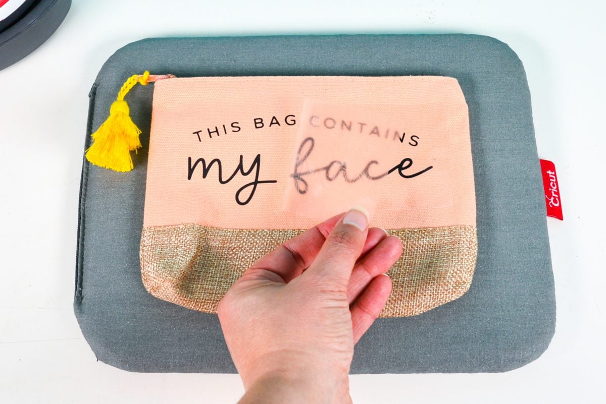 Hand peeling back the carrier sheet of the iron on vinyl on the makeup bag.