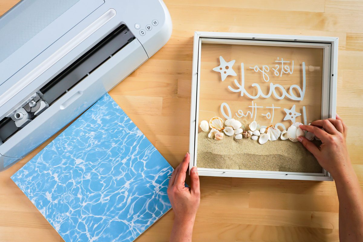 Hands filling shadow box with sand and shells.