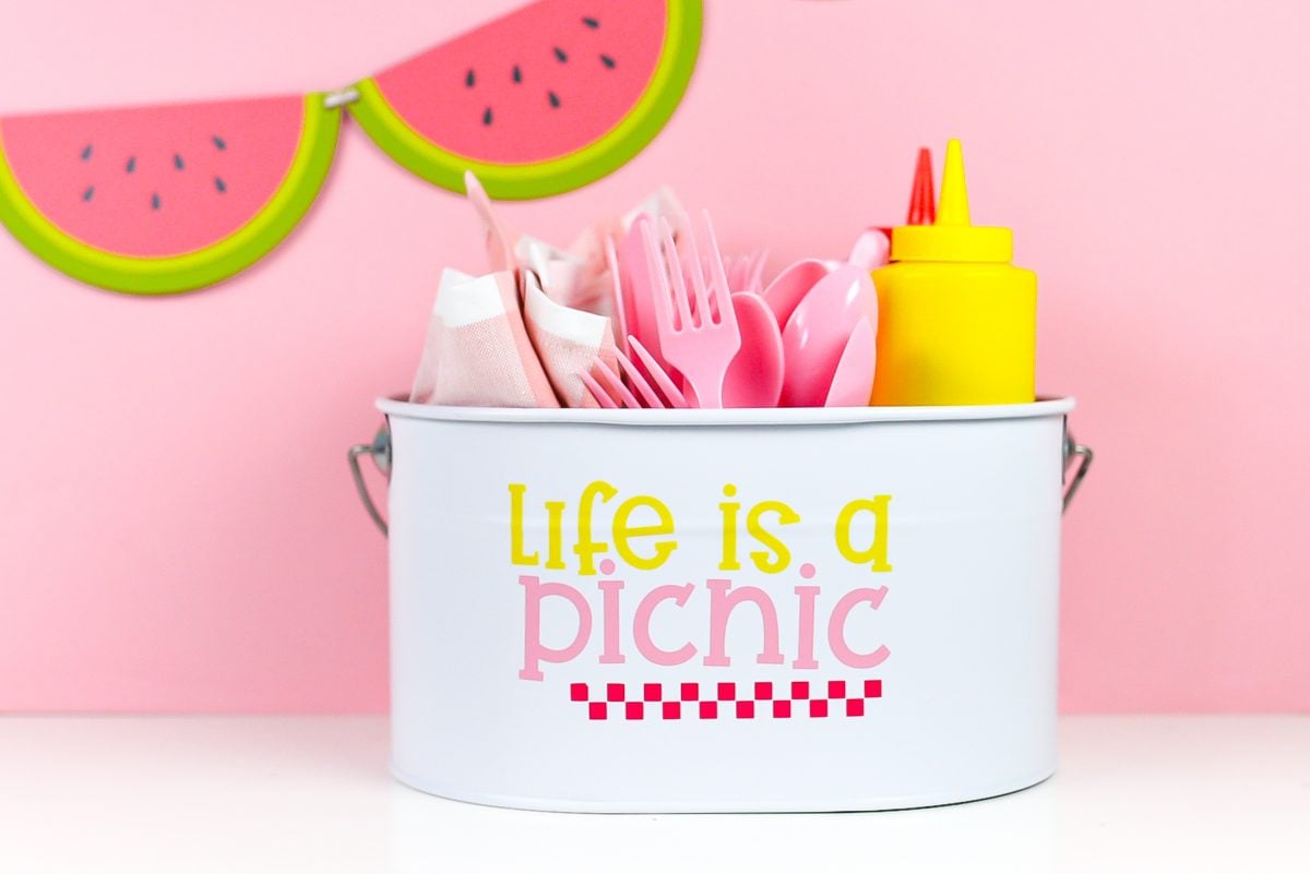 White picnic caddy with Life is a Picnic decal, filled with picnic supplies on a pink watermelon background.