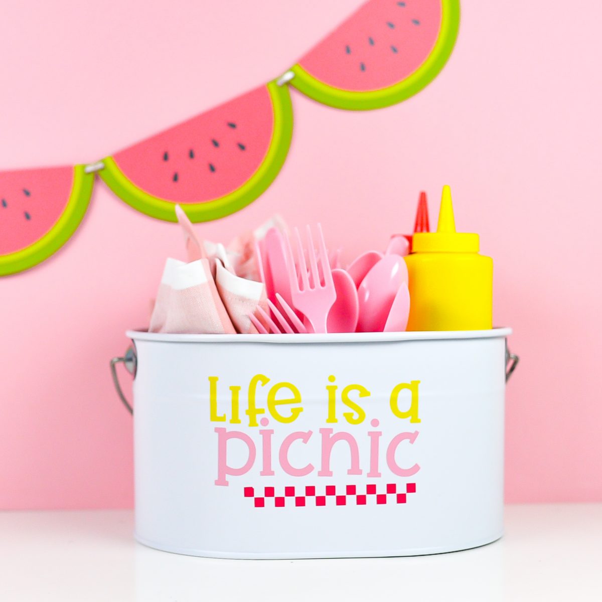 White picnic caddy with Life is a Picnic decal, filled with picnic supplies on a pink watermelon background.