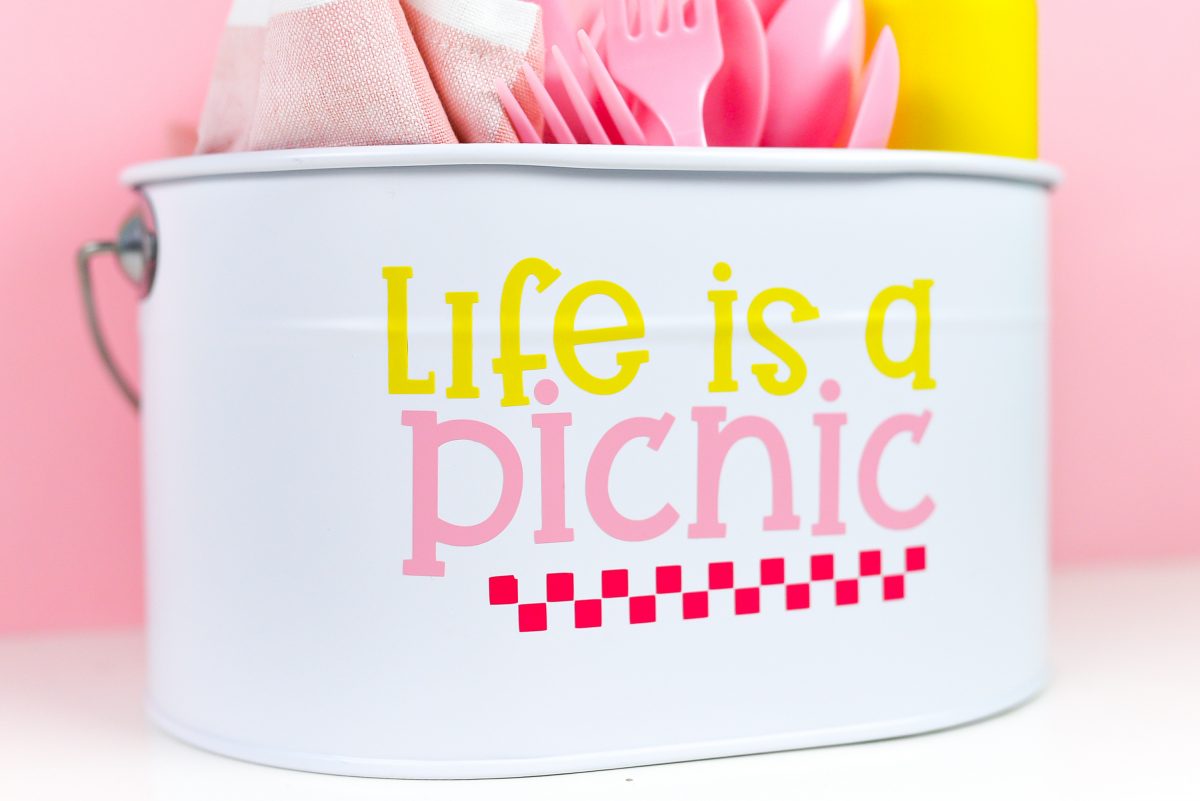 White picnic caddy with Life is a Picnic decal, filled with picnic supplies on a pink watermelon background.