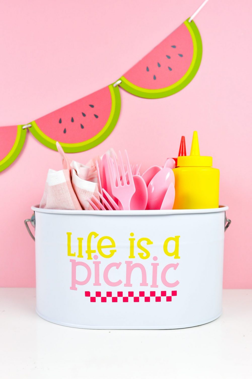 White picnic caddy with Life is a Picnic decal, filled with picnic supplies on a pink watermelon background.