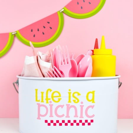 White picnic caddy with Life is a Picnic decal, filled with picnic supplies on a pink watermelon background.