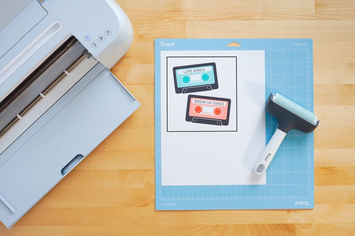 Overhead shot of printed mix tapes on a blue Cricut mat with a Cricut Maker 3 and brayer.