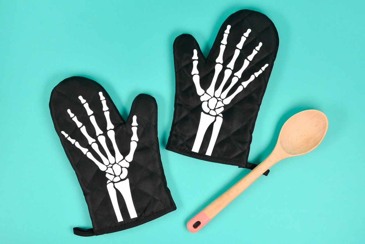 Skeleton oven mitts with spoon on teal background.