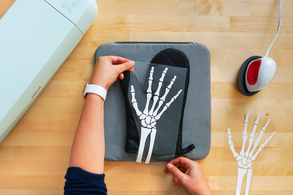 Hands placing iron on skeleton hand on black oven mitt with Cricut EasyPress Mini next to it.