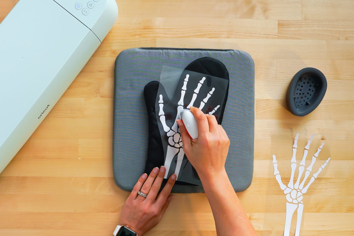 Hands using the EasyPress Mini to adhere the iron on vinyl skeleton hand to the oven mitt.