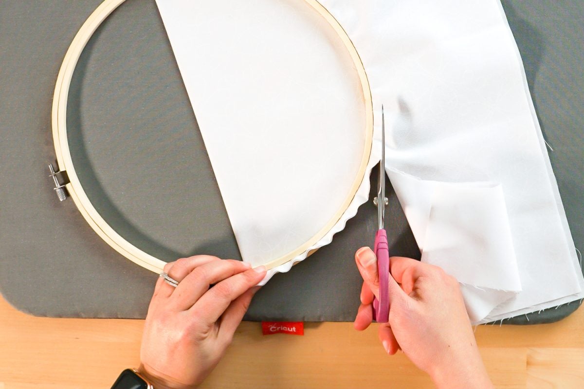 Hands using scissors to cut off excess fabric from the back of the hoop.