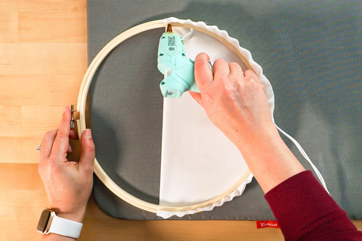 Hands using hot glue gun to secure fabric to back of embroidery hoop.