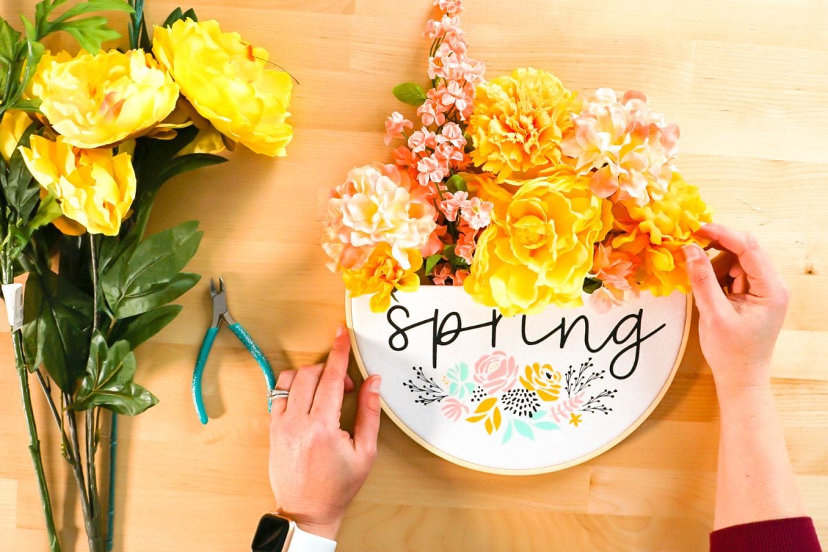 Hands adding final floral stems to embroidery hoop wreath. It's full of flowers.