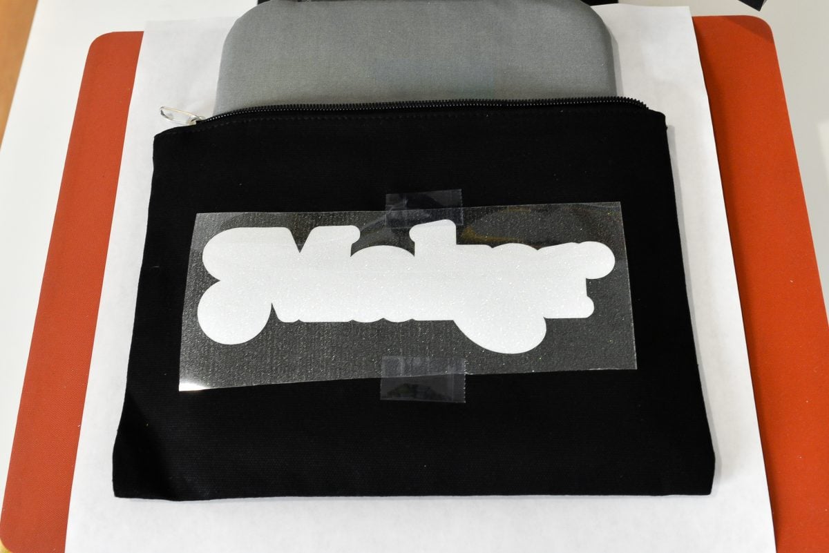 Glitter HTV image taped to black zippered pouch in heat press.