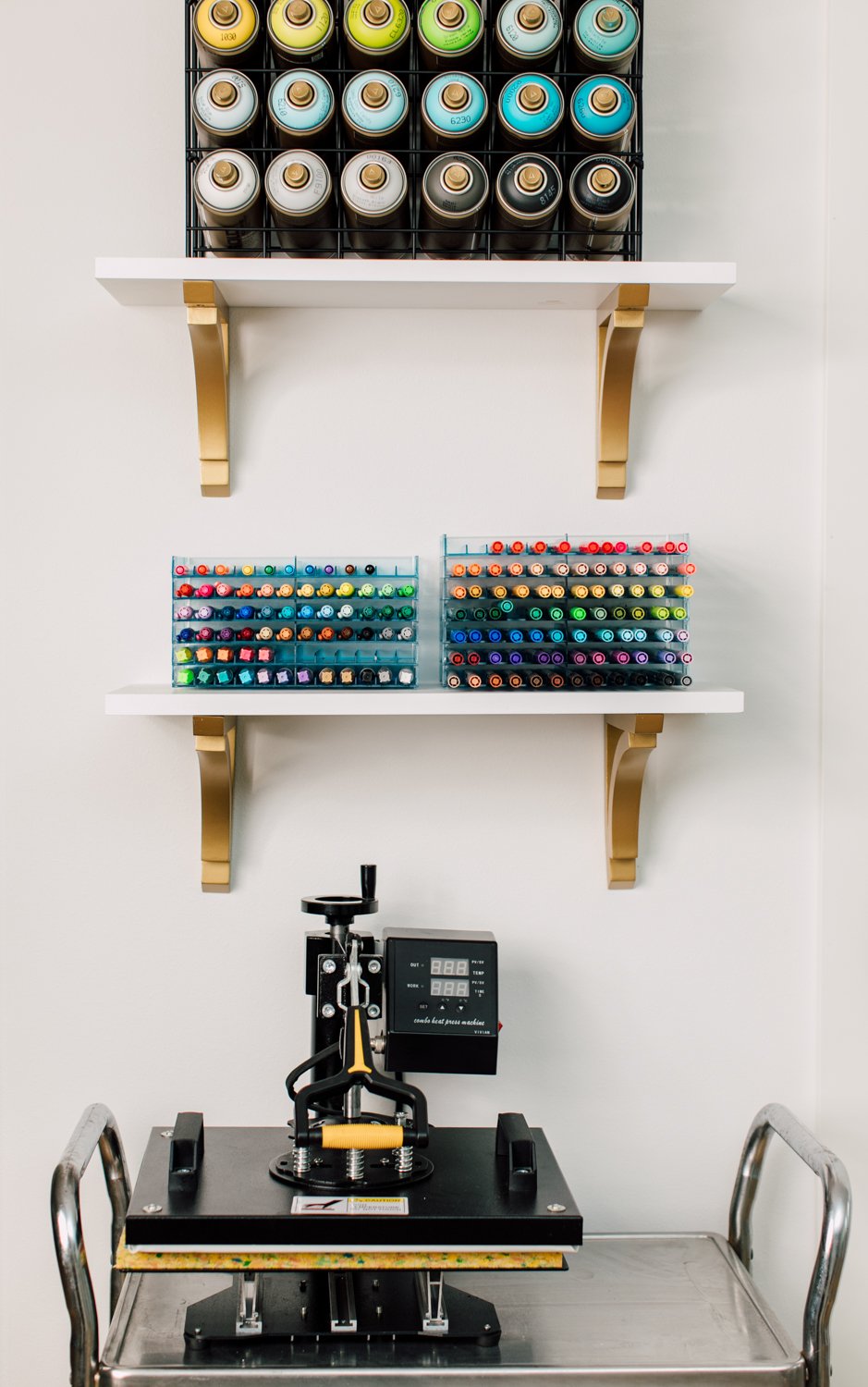 Heat press on cart, with shelves of pens and spray paint above. 