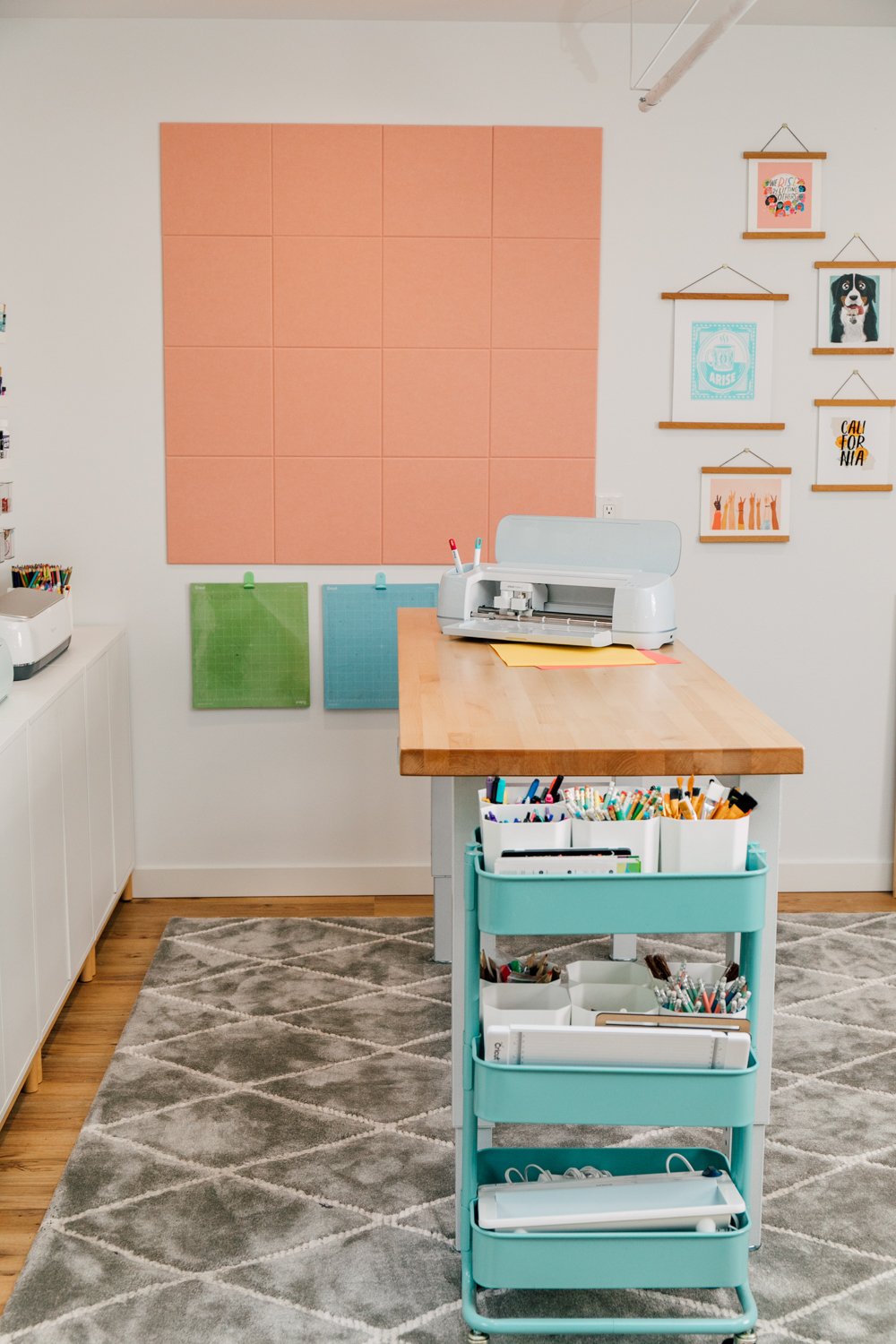 Side view of worktable with Cricut on top, rolling blue cart with pens, and felt squares on the wall.