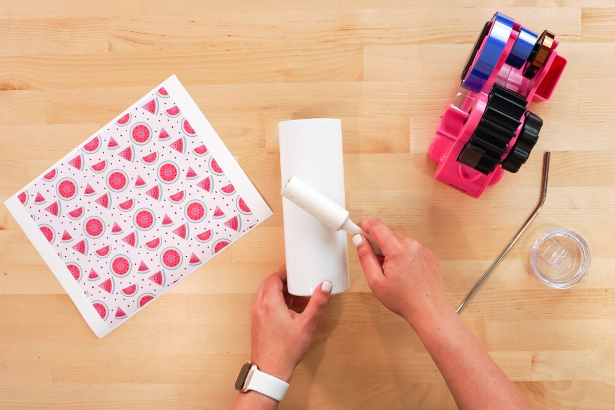 Hands cleaning a blank tumbler with a lint roller.