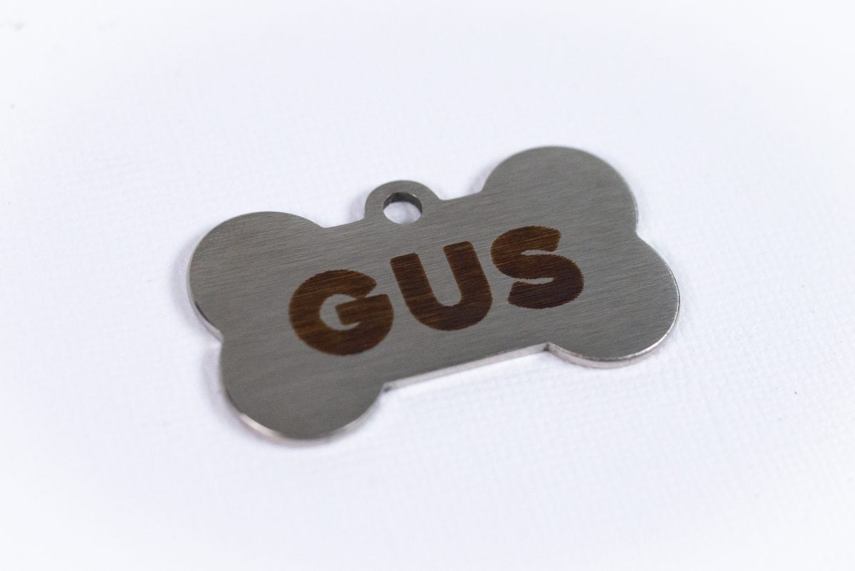 Image of a piece of metal in the shape of a dog bone with the name Gus engraved on it