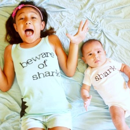 A young girl wearing a tank top with the words Beware of Shark on it and a baby wearing a white onesie with the word Shark on it