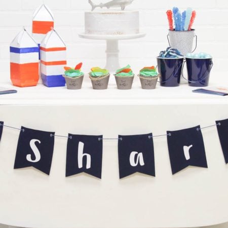 A table set up with a cake and other party items and a banner across the front of the table that says Shark