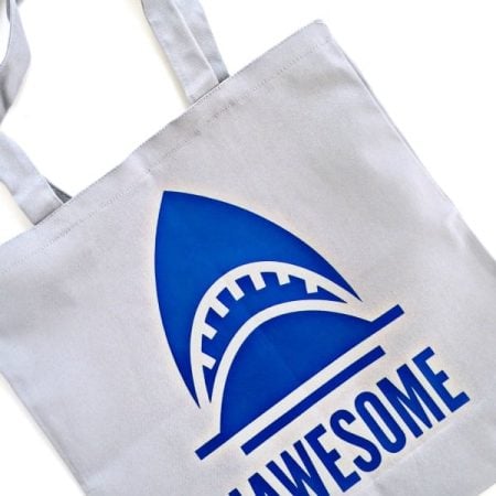 A white tote bag with an image of a blue shark on it and the word Jawesome
