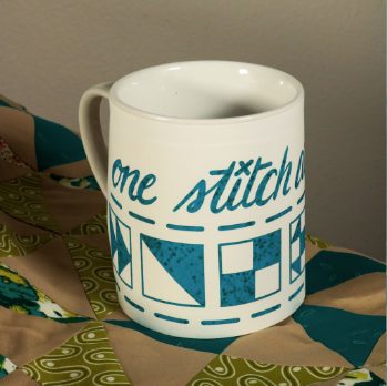 White coffee mug decorated in a teal blue quilt design that says One Stitch at a Time