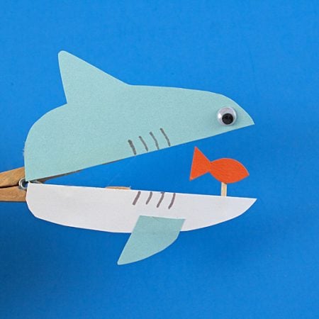 A hand holding a clothespin that has an image glued to it of a paper cut out shark with a small fish inside of its mouth