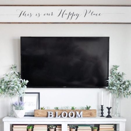 A large wooden sign that says This is our Happy Place hanging above a large TV and a TV stand filled with decor
